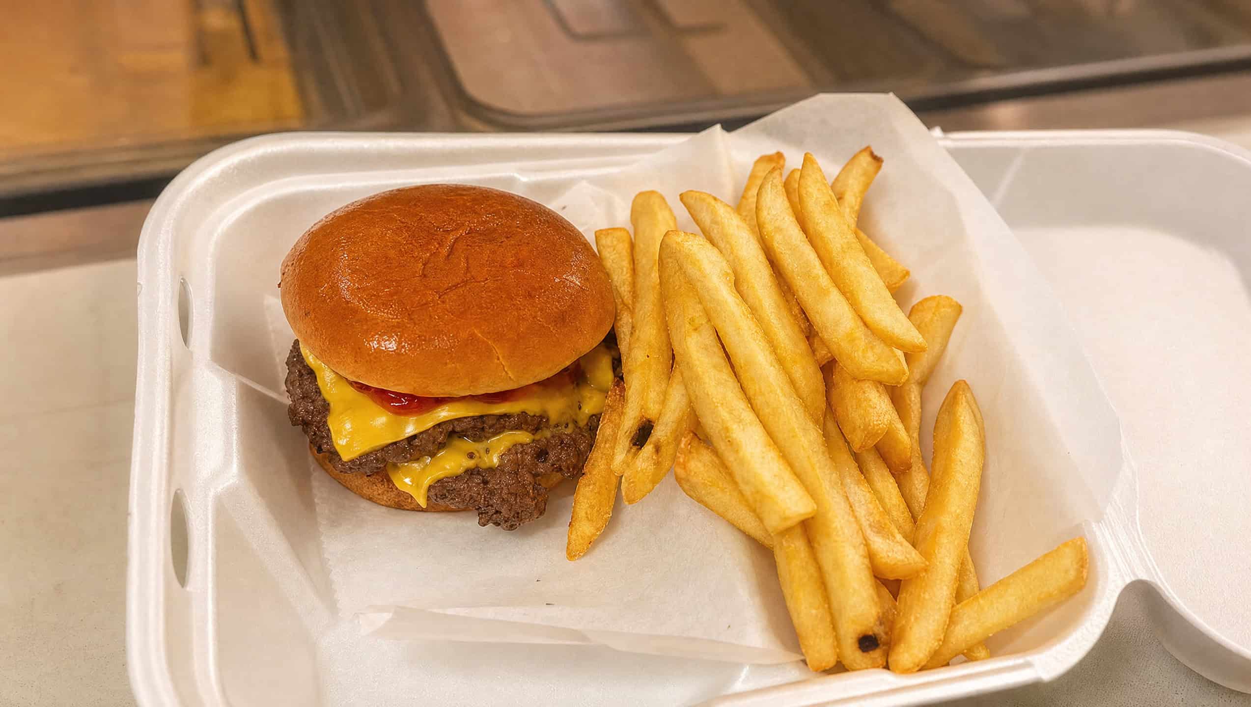 A cheeseburger with melted American cheese and ketchup on a toasted brioche bun, served with golden French fries in a takeout container.