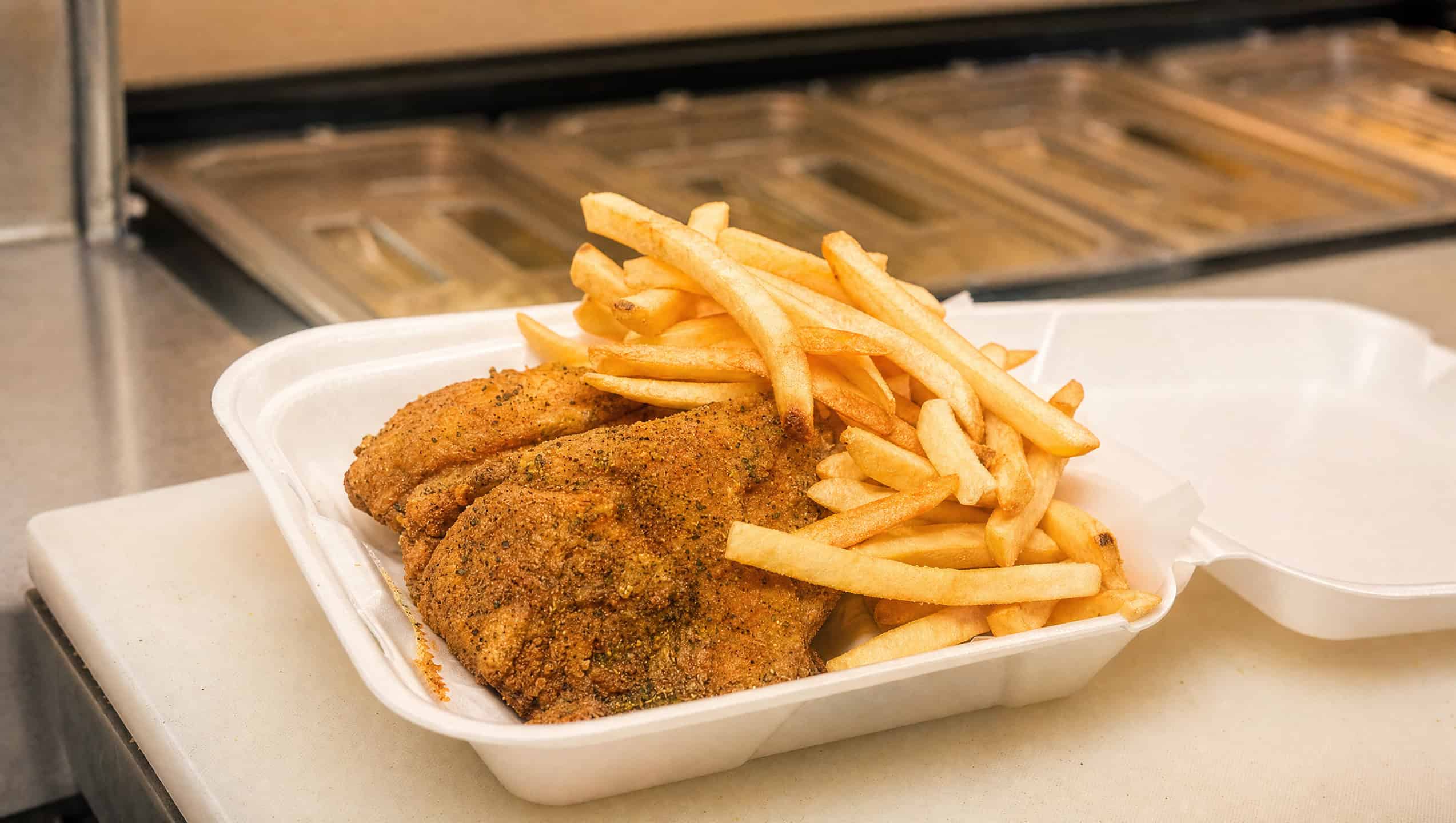 Golden, seasoned deep-fried catfish fillets served with crispy French fries in a takeout container.